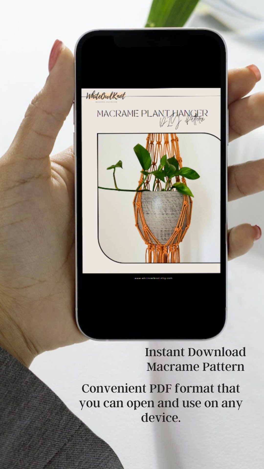 Hand holding a smartphone displaying a macrame plant hanger pattern with text about instant download.
