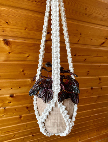 White macrame plant hanger with a plant against a wooden wall.