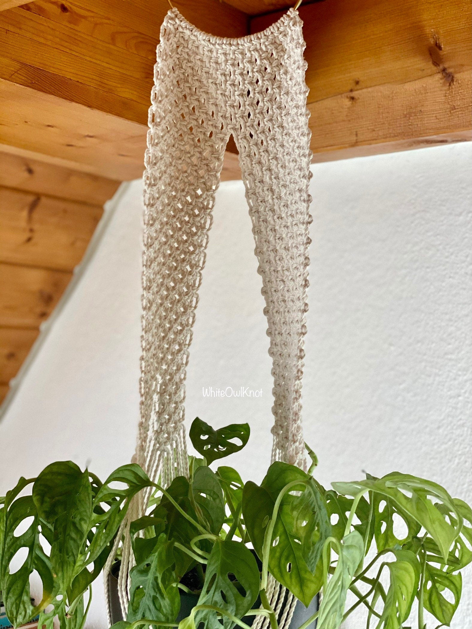 Beige crochet pants hanging on a plant against a wooden ceiling and white wall.