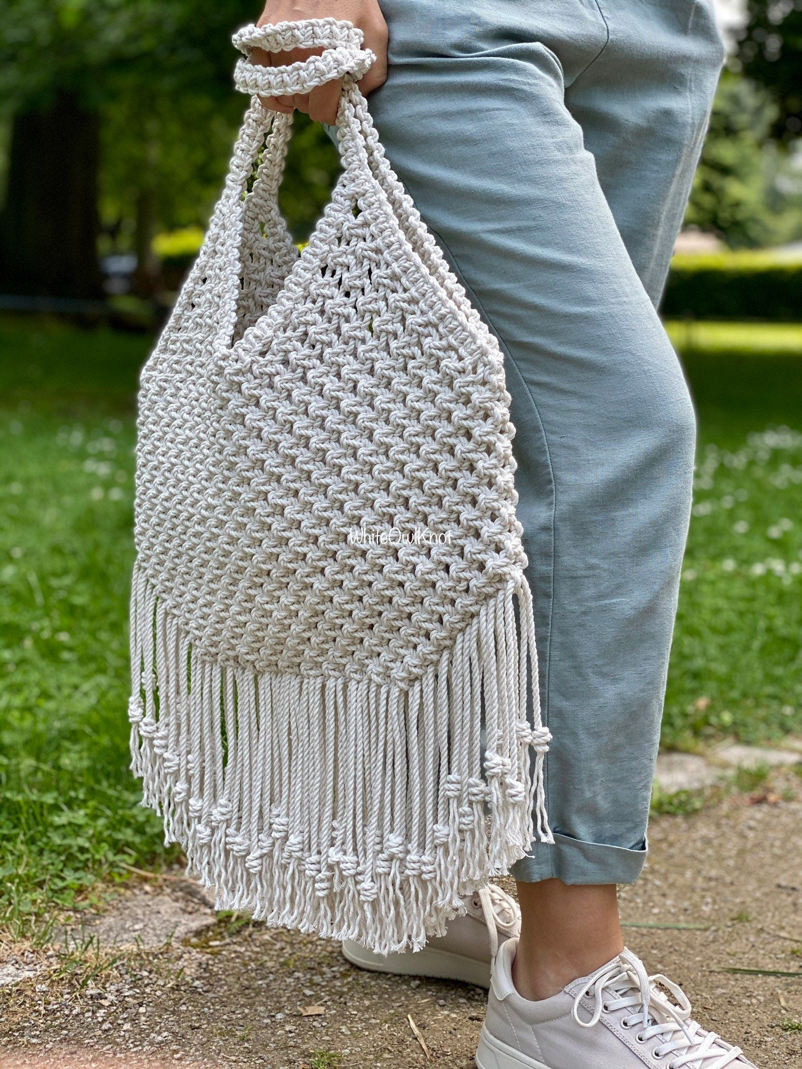 Person holding a white macrame bag with fringes outdoors on a grassy area.