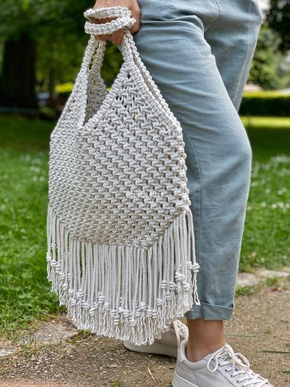 Person holding a white macrame bag with fringes outdoors on a grassy area.
