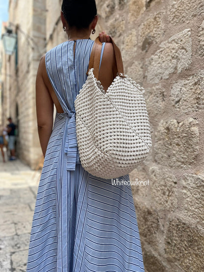 Woman in a blue dress carrying a white woven macrame bag against a stone wall.
