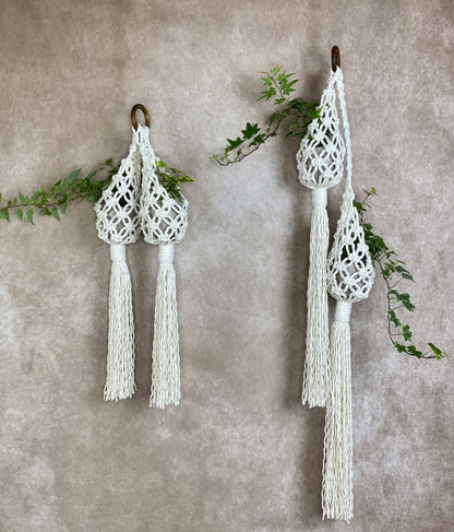 Two white macrame plant hangers with tassels on a textured wall.