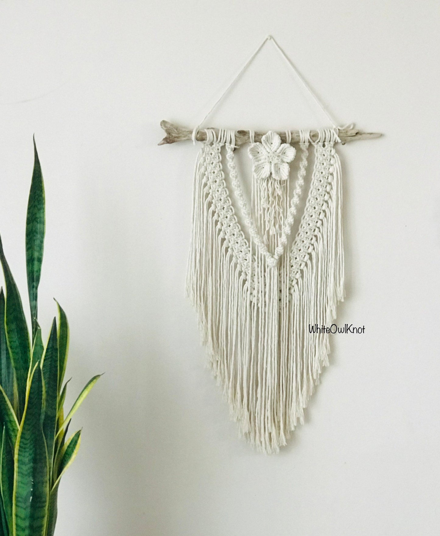 Macrame wall hanging with floral element on a white wall, featuring the brand 'White Owl Knot'.