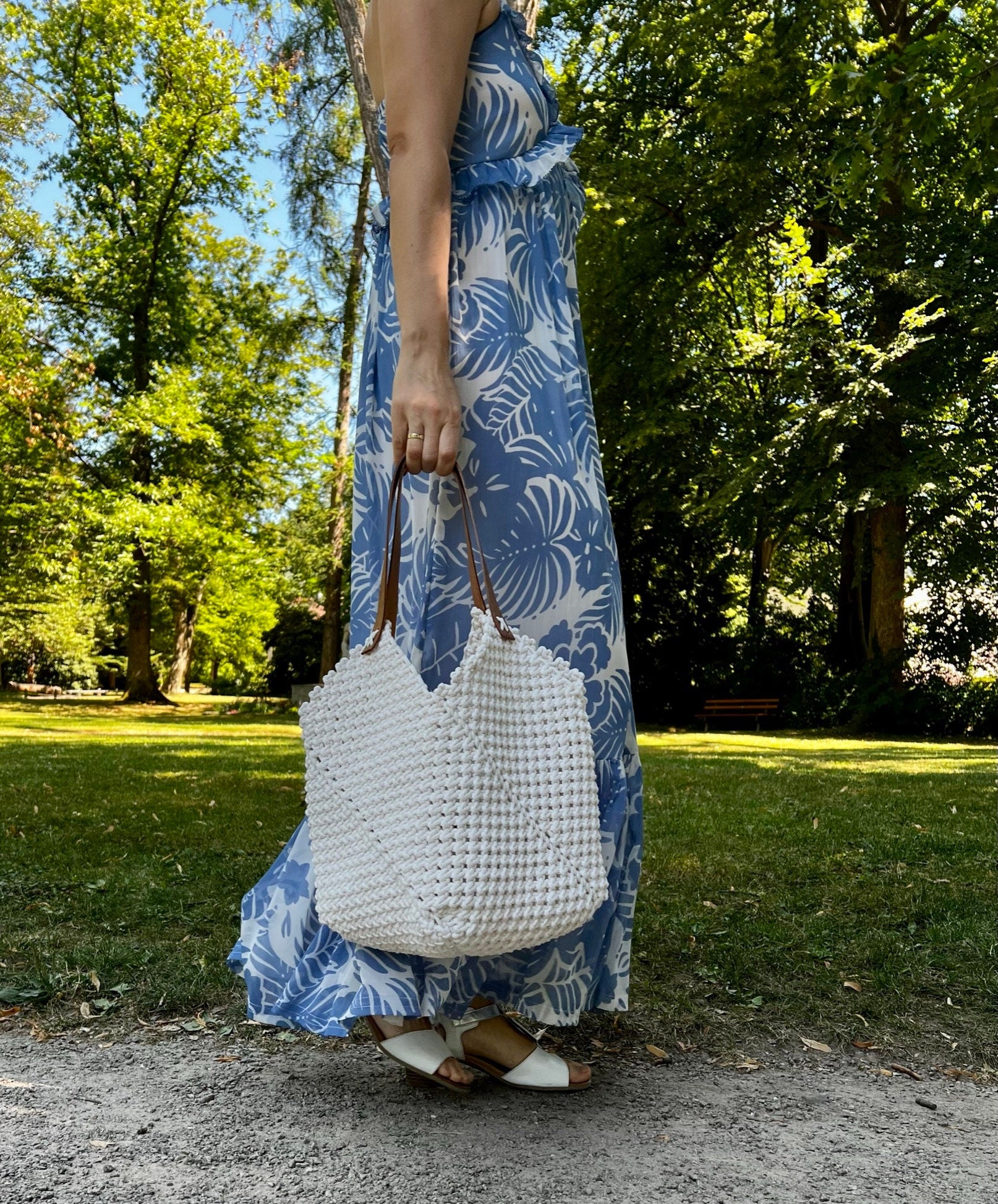 Person wearing a blue floral dress holding a white woven macrame bag in a park setting
