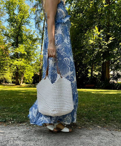 Person wearing a blue floral dress holding a white woven macrame bag in a park setting