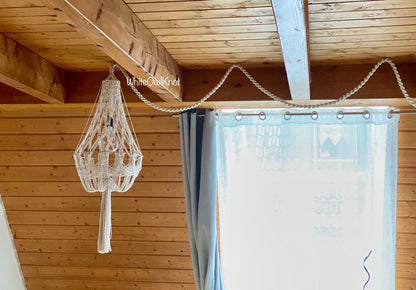 Decorative macrame light fixture hanging from a wooden ceiling with a curtain in the background.