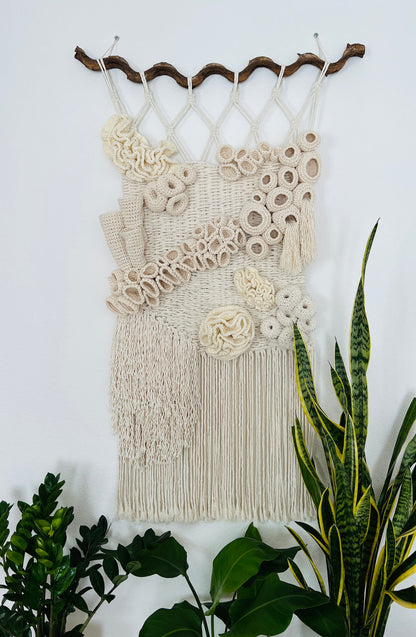 Macrame wall hanging with coral reef accents on a white wall with plants below.