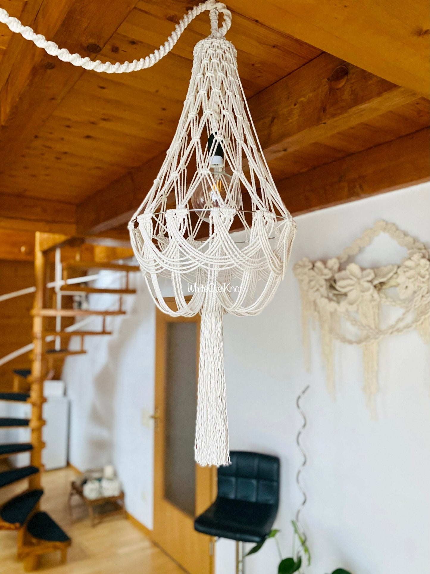 Macrame chandelier hanging in a room with wooden ceiling and staircase.