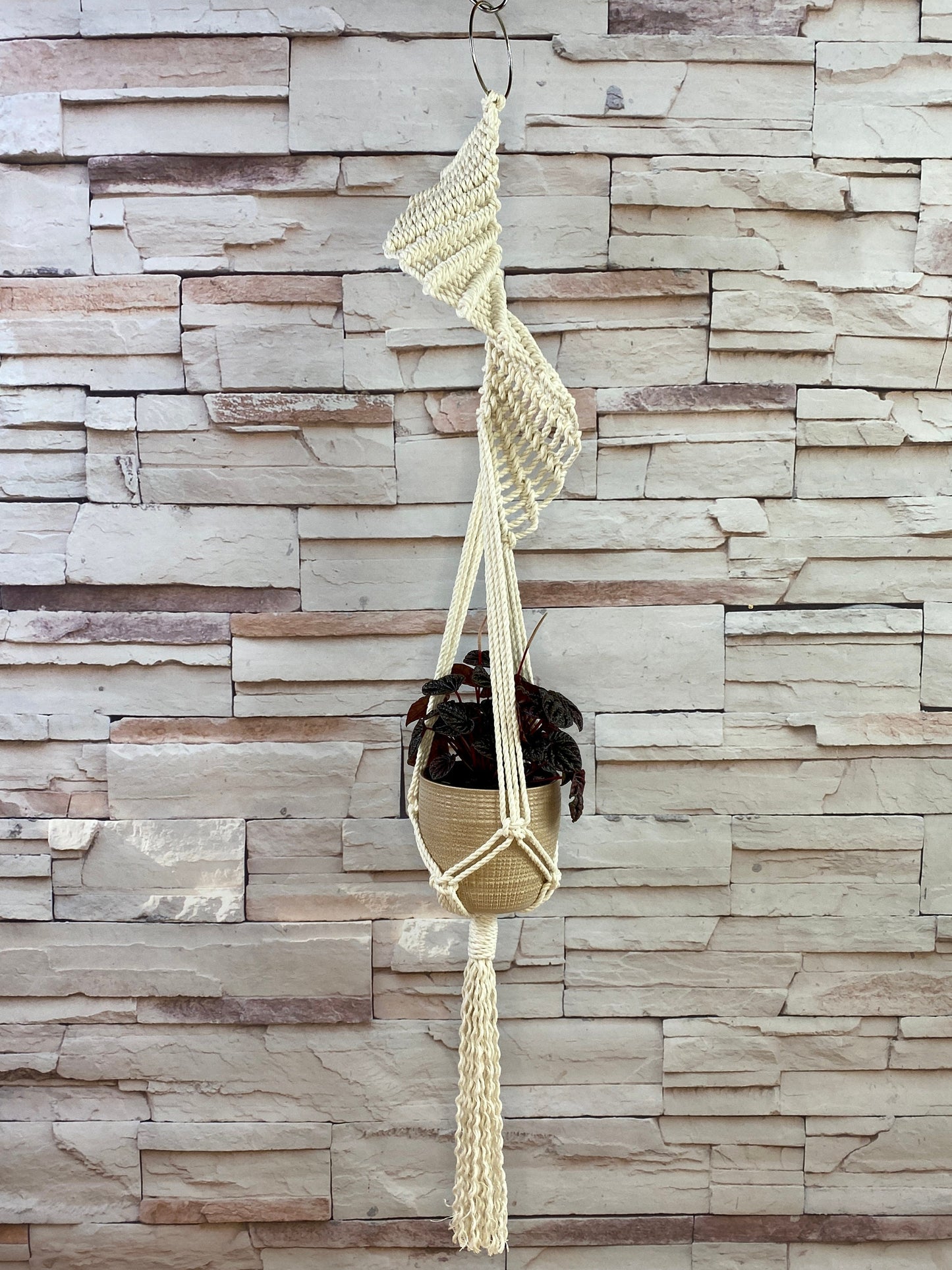 Decorative plant holder with tassels hanging against a stone wall.