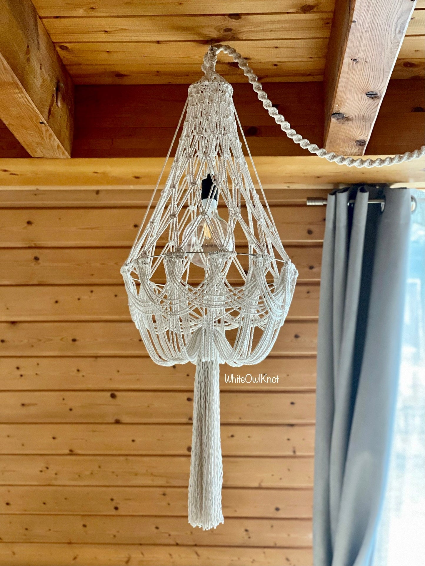 Macrame chandelier hanging from a wooden ceiling with a curtain in the background.