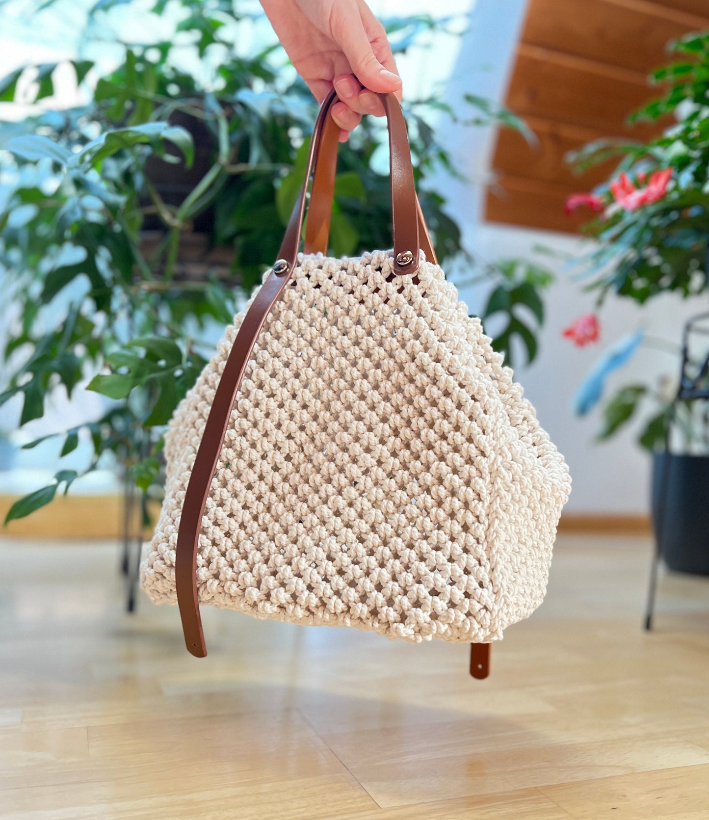 Beige woven handbag with brown handles held by a hand indoors with plants in the background