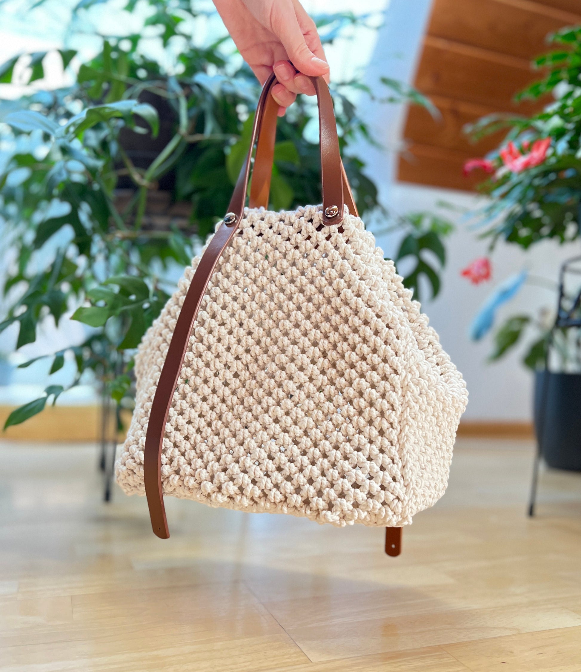 Beige woven handbag with brown handles held by a hand indoors with plants in the background