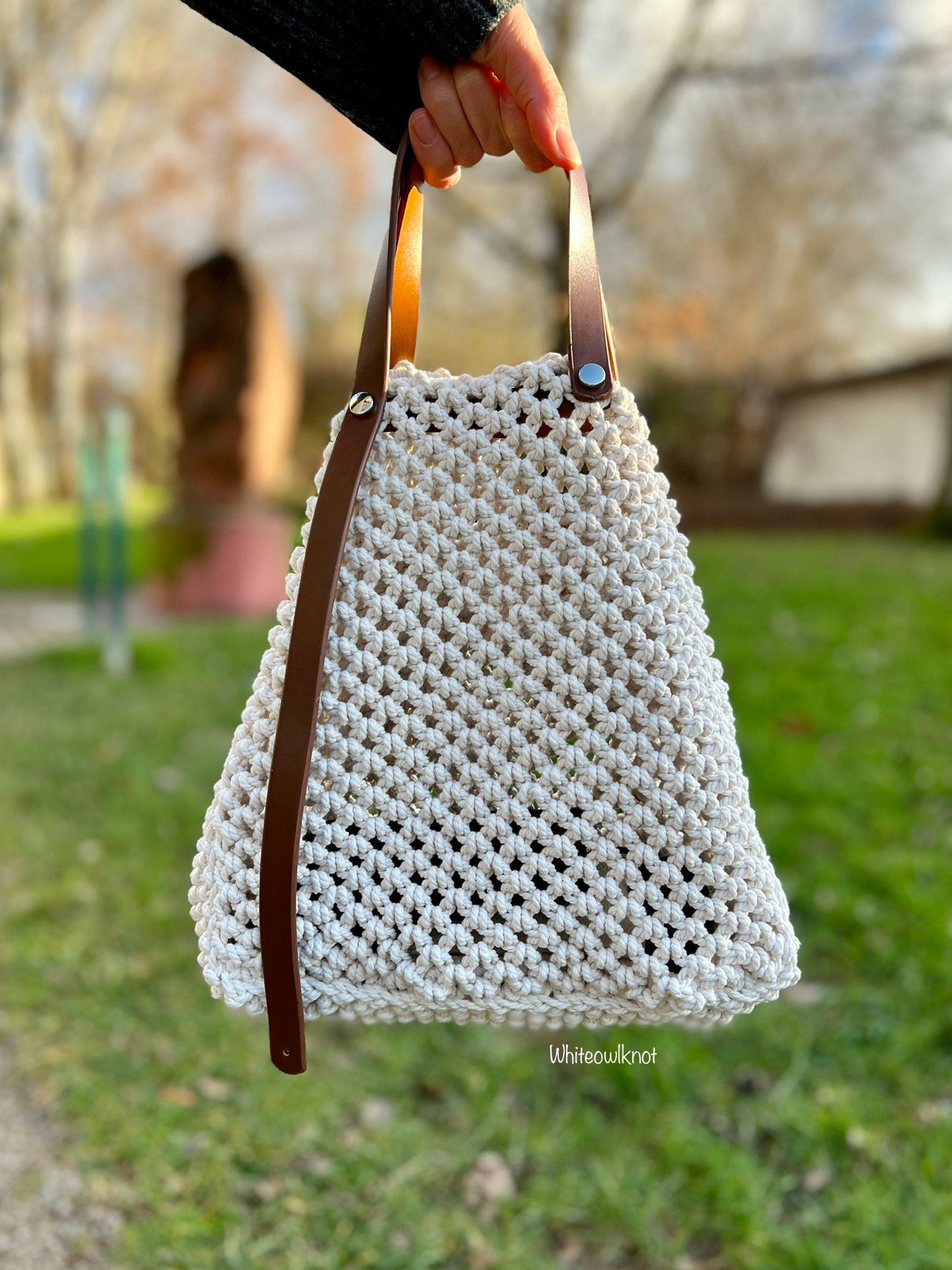 White woven macrame handbag with brown leather straps held by a person outdoors.