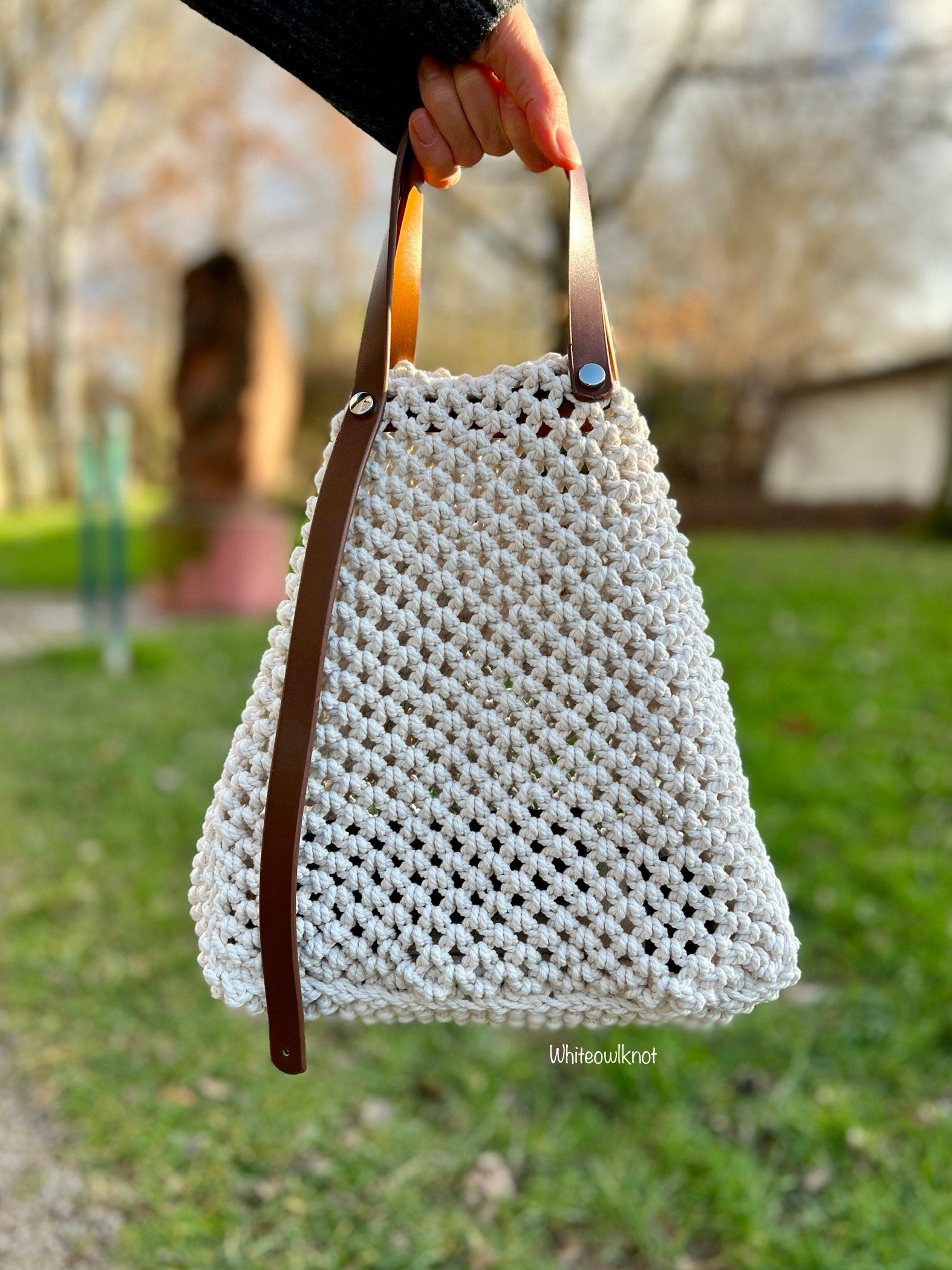 White woven macrame handbag with brown leather straps held by a person outdoors.