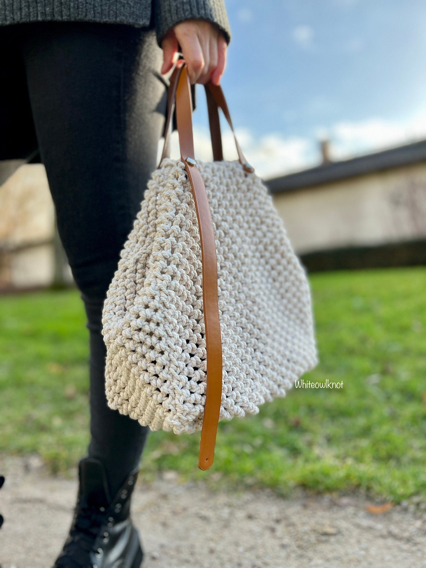 Person holding a beige woven macrame handbag with a brown strap outdoors.