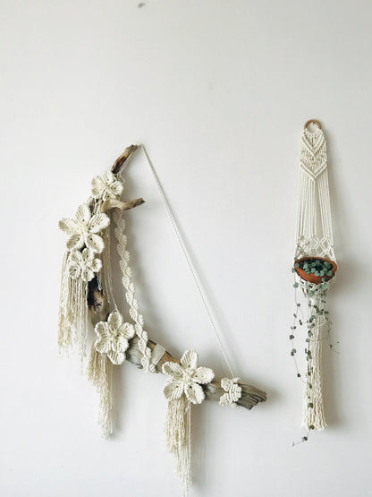 Macrame wall hanging with floral decorations and a small container on a white background