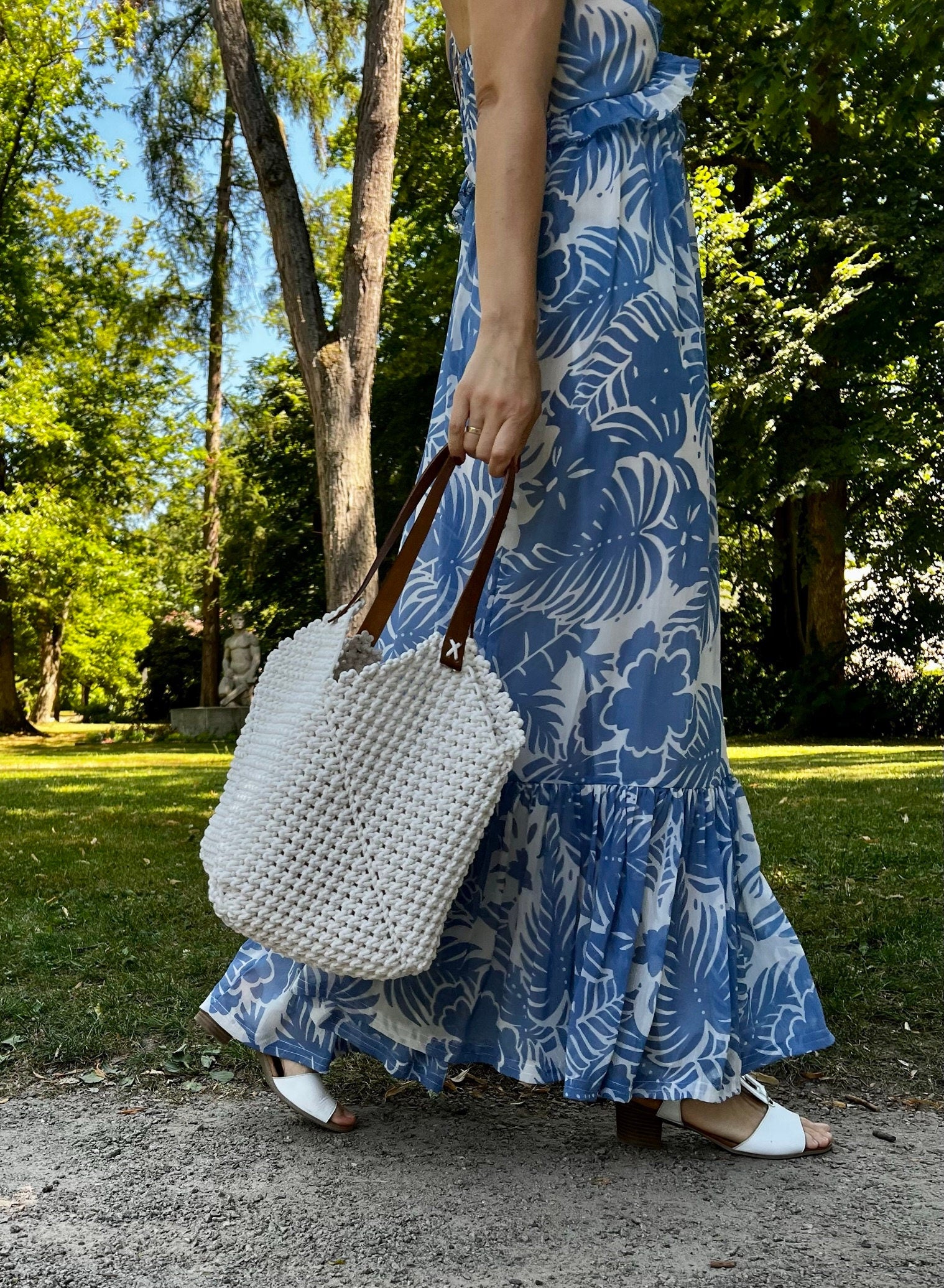 Person wearing a blue floral dress holding a white textured handbag in a park setting.