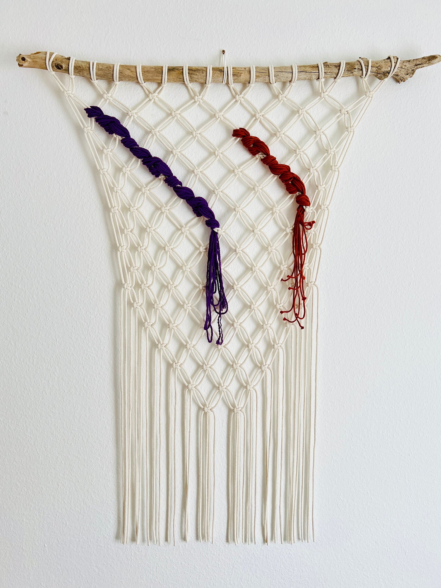 Macrame wall hanging with white, purple, and red strings on a plain background