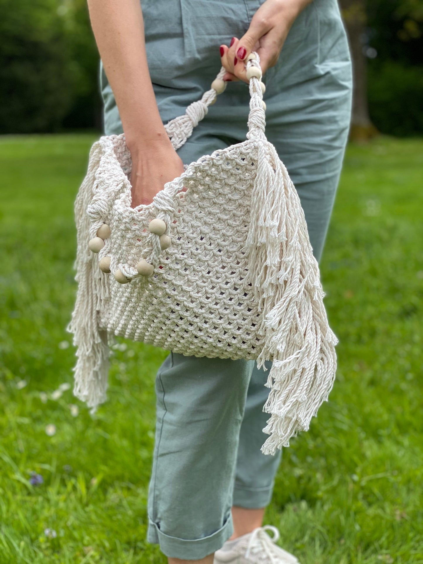Person holding a white macrame bag with tassels outdoors on grass