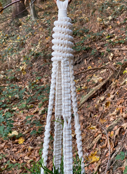 White macrame plant hanger hanging in a natural outdoor setting with trees and leaves.