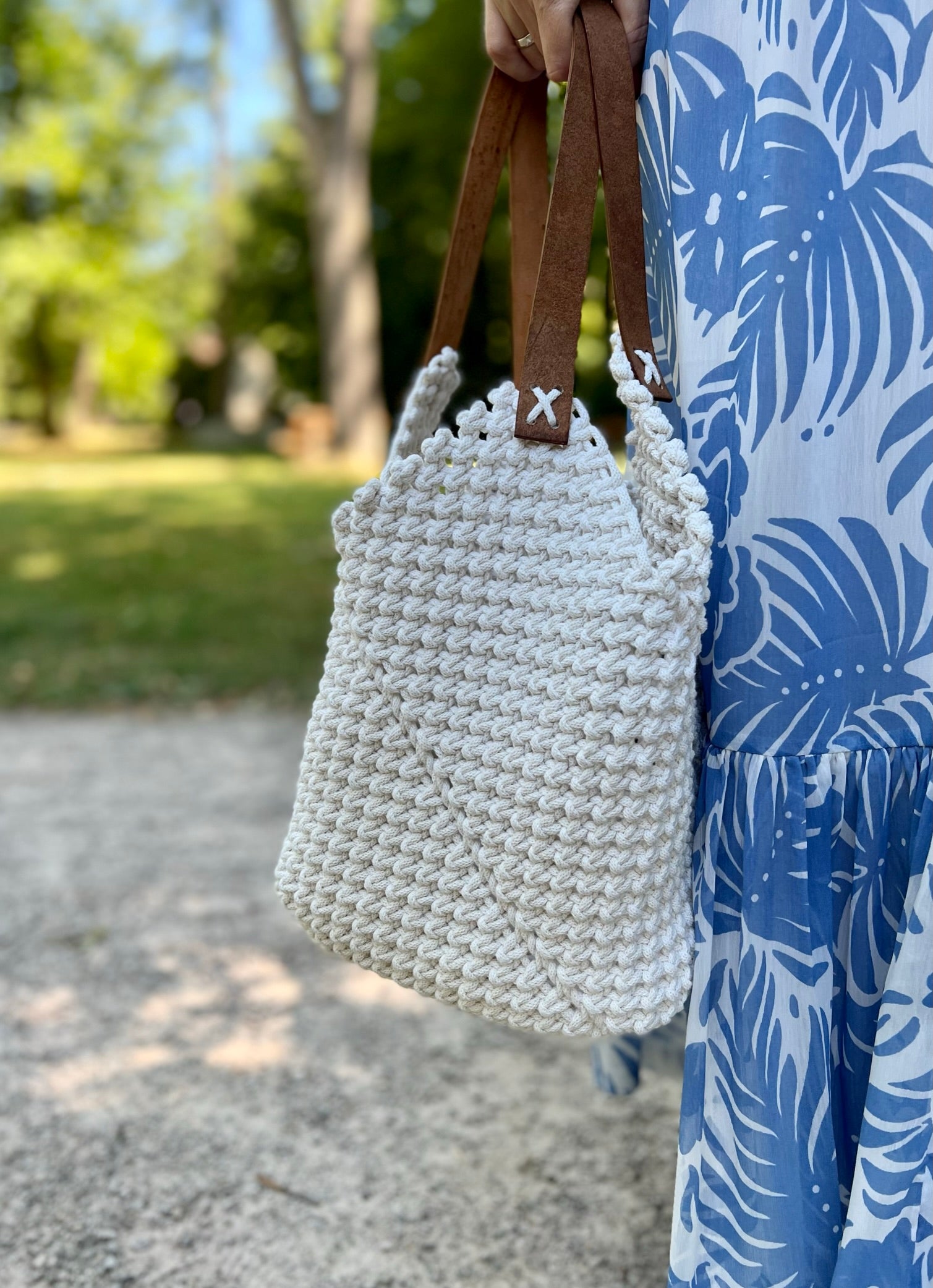 White woven handbag with brown leather straps held by a person wearing a blue and white patterned dress outdoors.