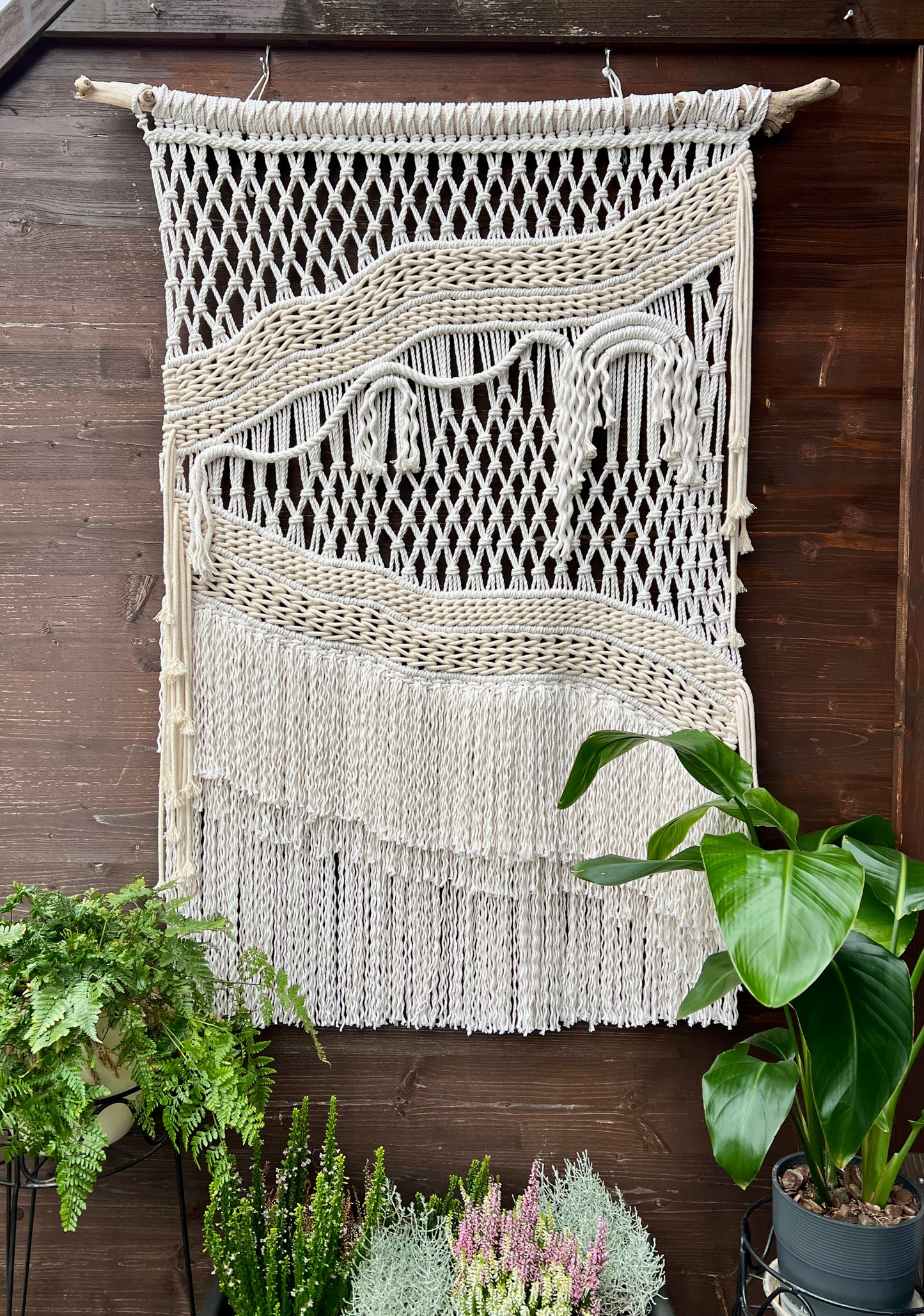 Macrame wall hanging on a wooden wall with plants below