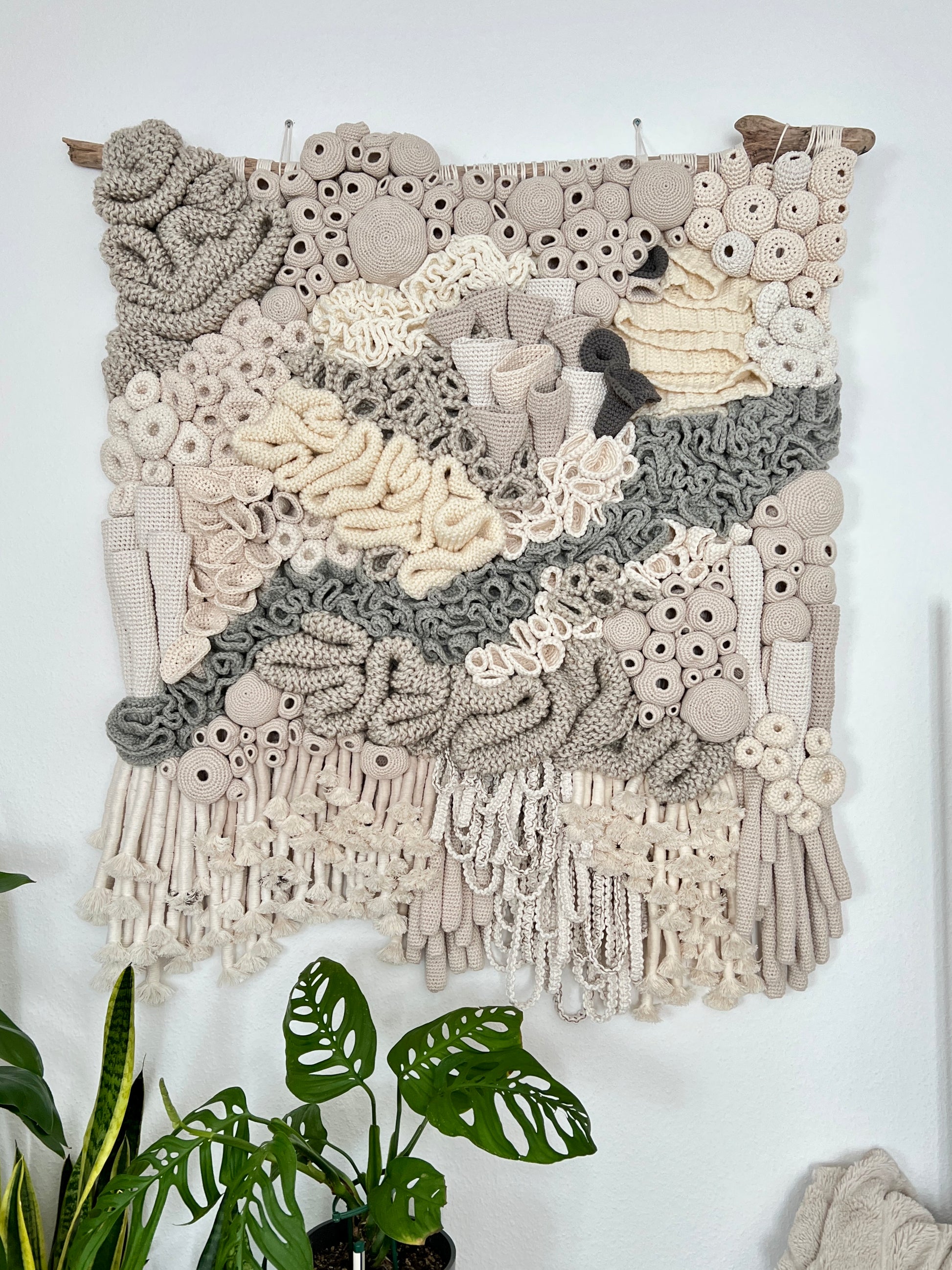 Textile art piece with intricate patterns on a white wall, featuring plants below.