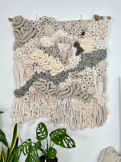 Textile art piece with intricate patterns on a white wall, featuring plants below.