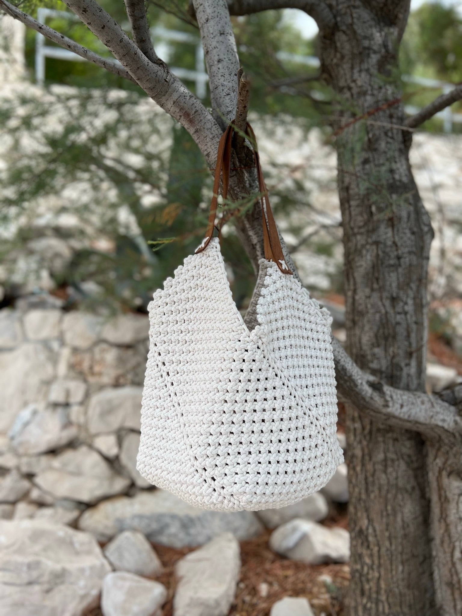 White woven bag hanging from a tree in a natural setting with rocks and greenery.