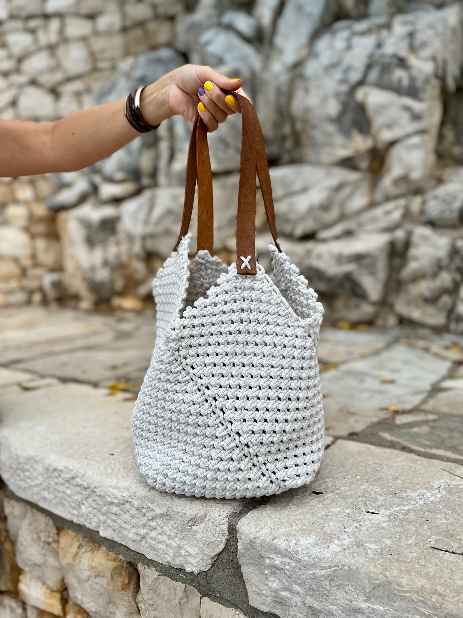 White woven bag with brown handles held by a hand against a stone wall background