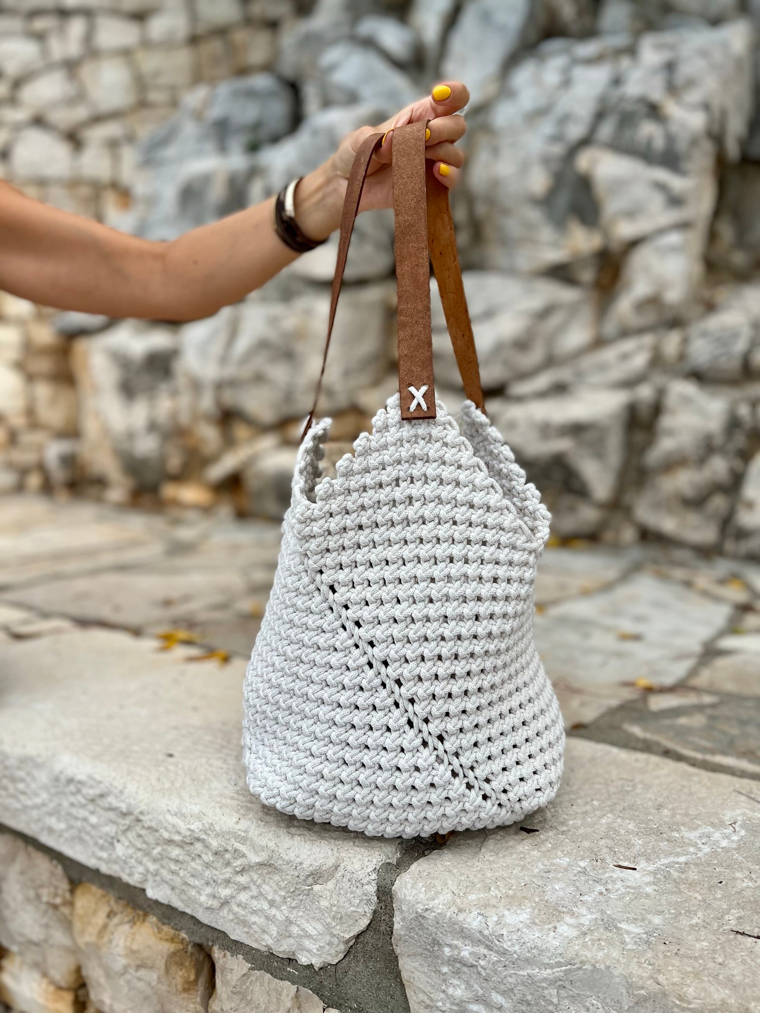 White woven handbag with brown leather straps held by a person against a stone wall.