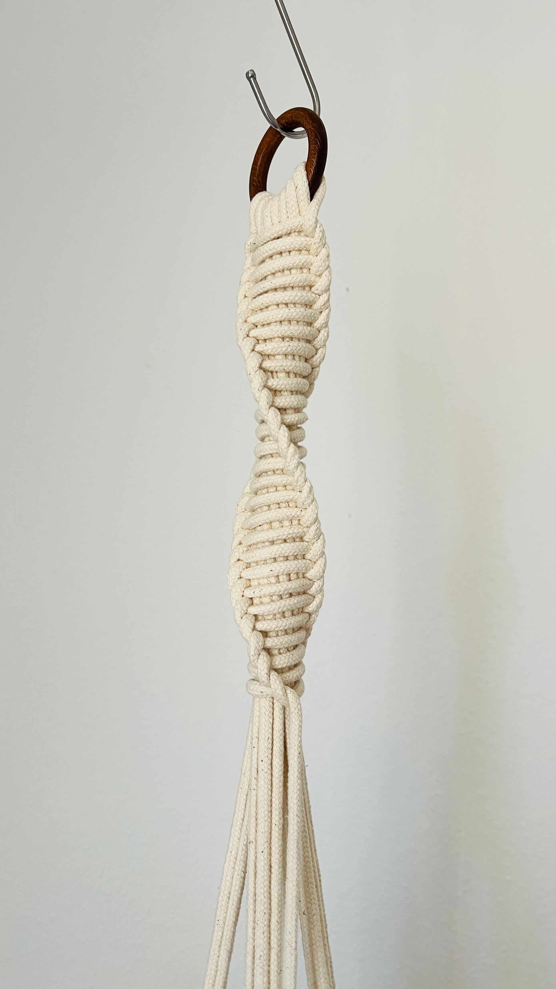White macrame plant hanger hanging on a hook against a plain background