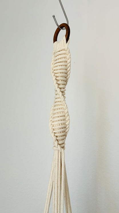 White macrame plant hanger hanging on a hook against a plain background