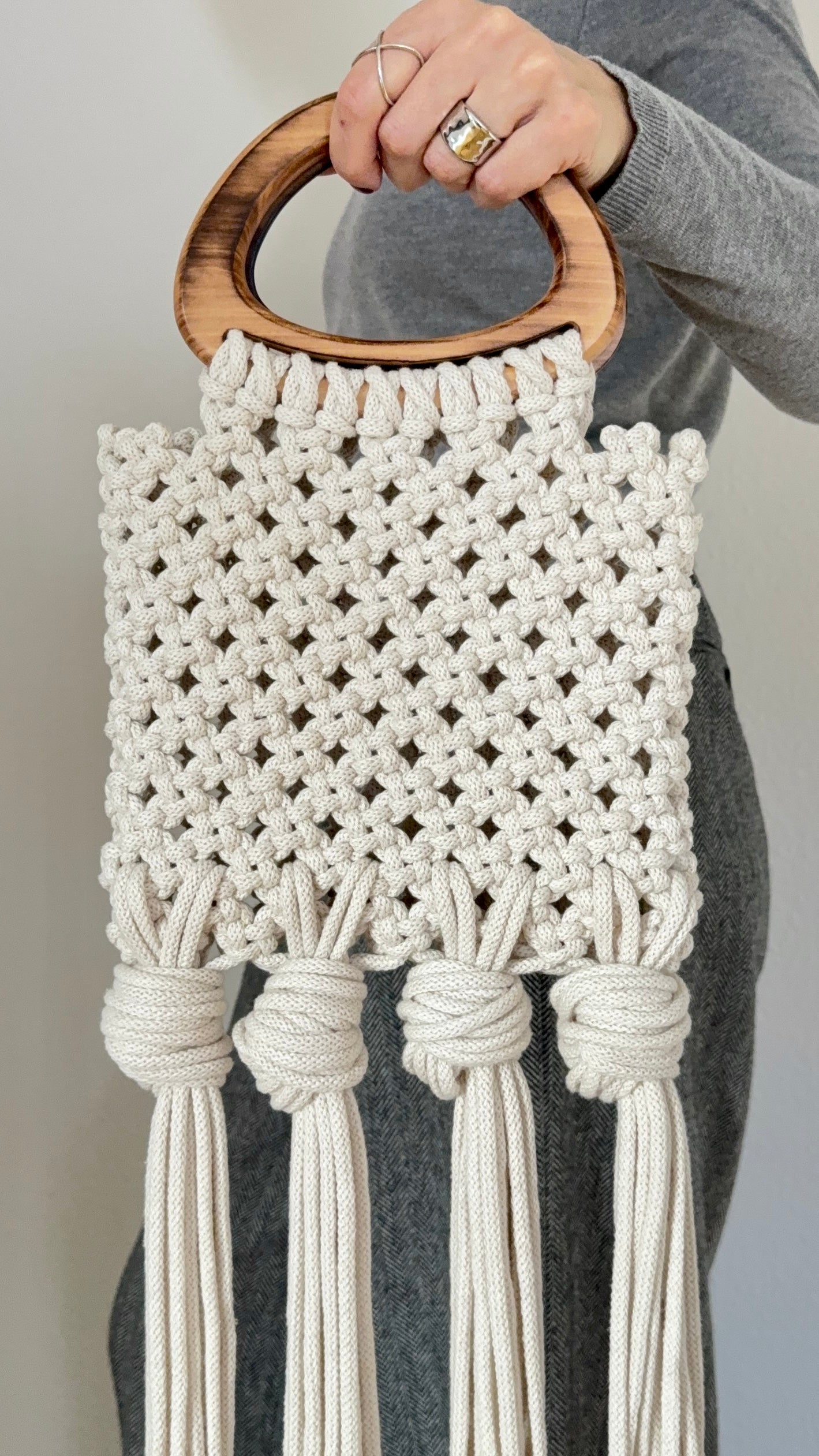 Macrame wall hanging with wooden handle held by a person against a neutral background