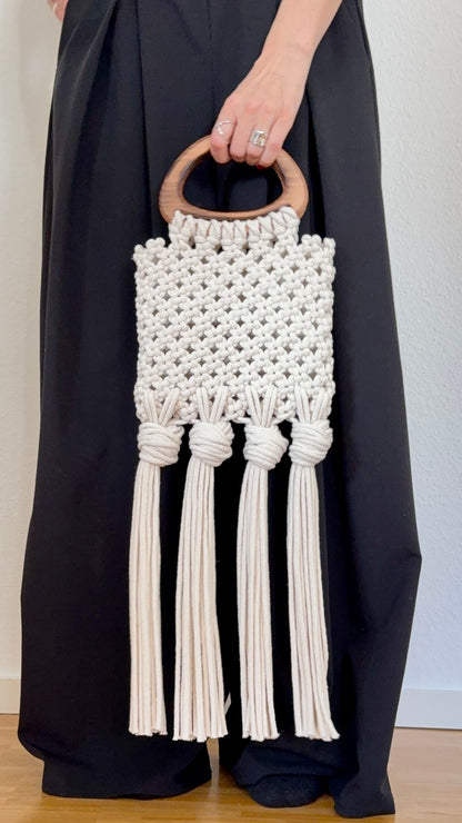 Handmade macrame bag with wooden handle held by a person wearing a black outfit.