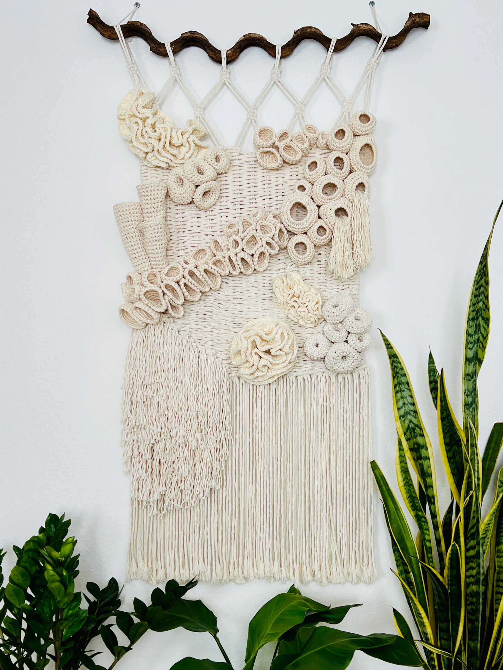 Macrame wall hanging with floral patterns on a white wall with plants around