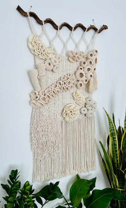 Macrame wall hanging with decorative elements on a white wall with plants around.