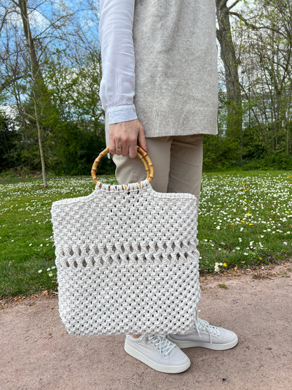 Person holding a white macrame tote bag outdoors on a path with greenery.