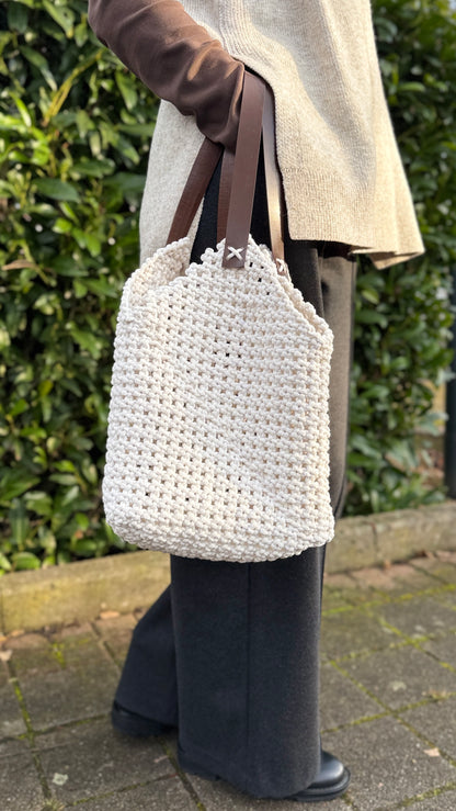 Person holding a white crochet bag with brown straps against a green hedge background