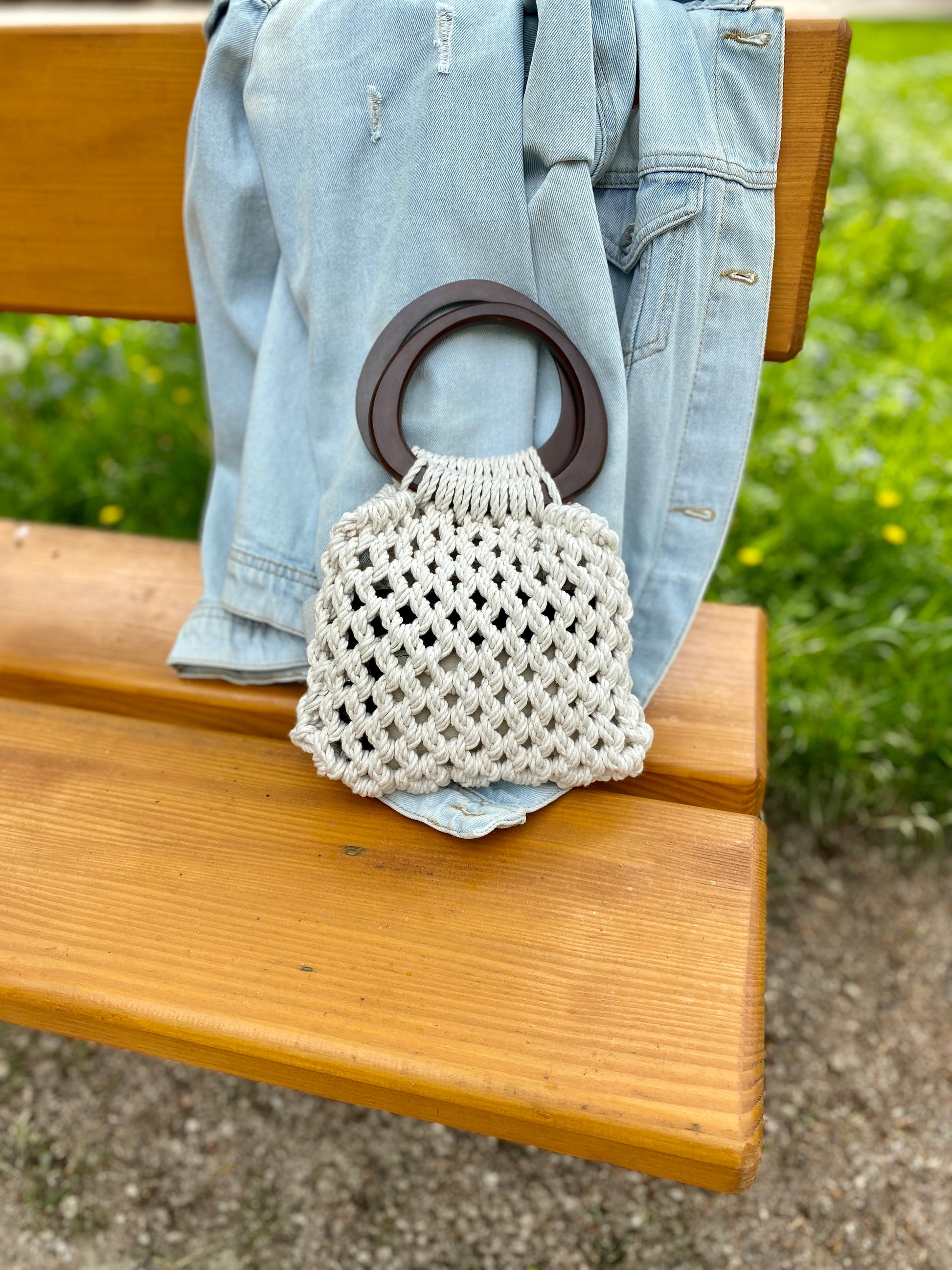 Macrame handbag with wooden handle on a denim jacket over a wooden bench outdoors.