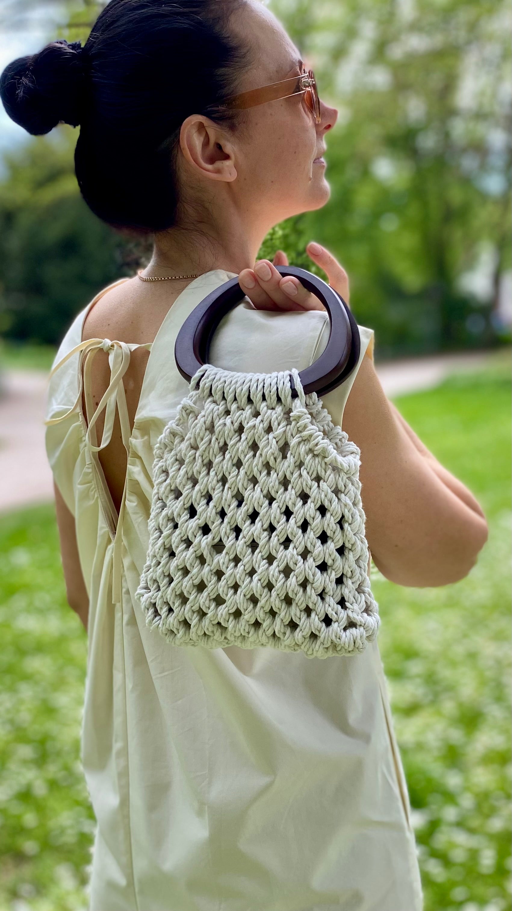 Woman holding a white macrame bag with wooden handle outdoors.