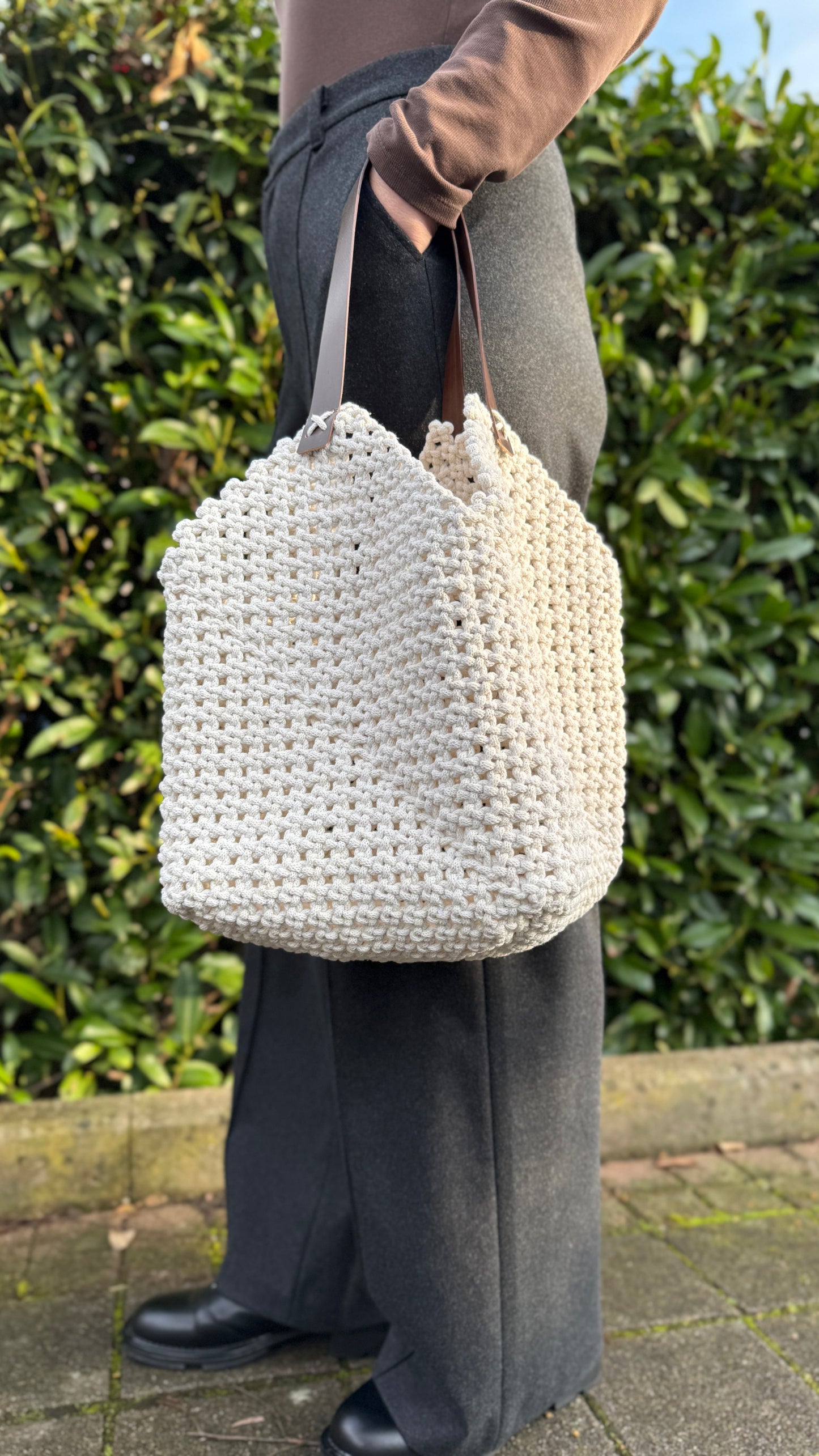 Person holding a white woven bag against a green hedge background