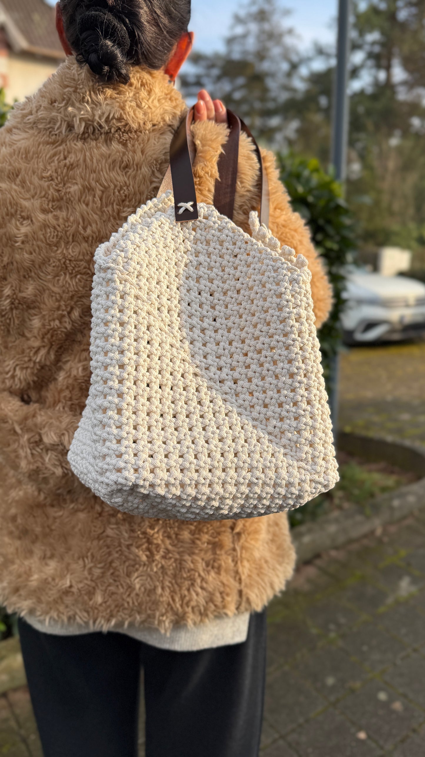 Person wearing a beige fluffy coat holding a beige macrame bag outdoors.