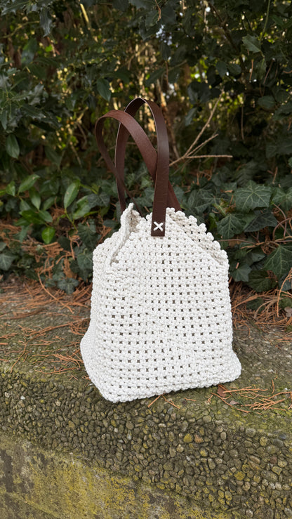White macrame bag with brown leather straps on a stone surface with greenery in the background