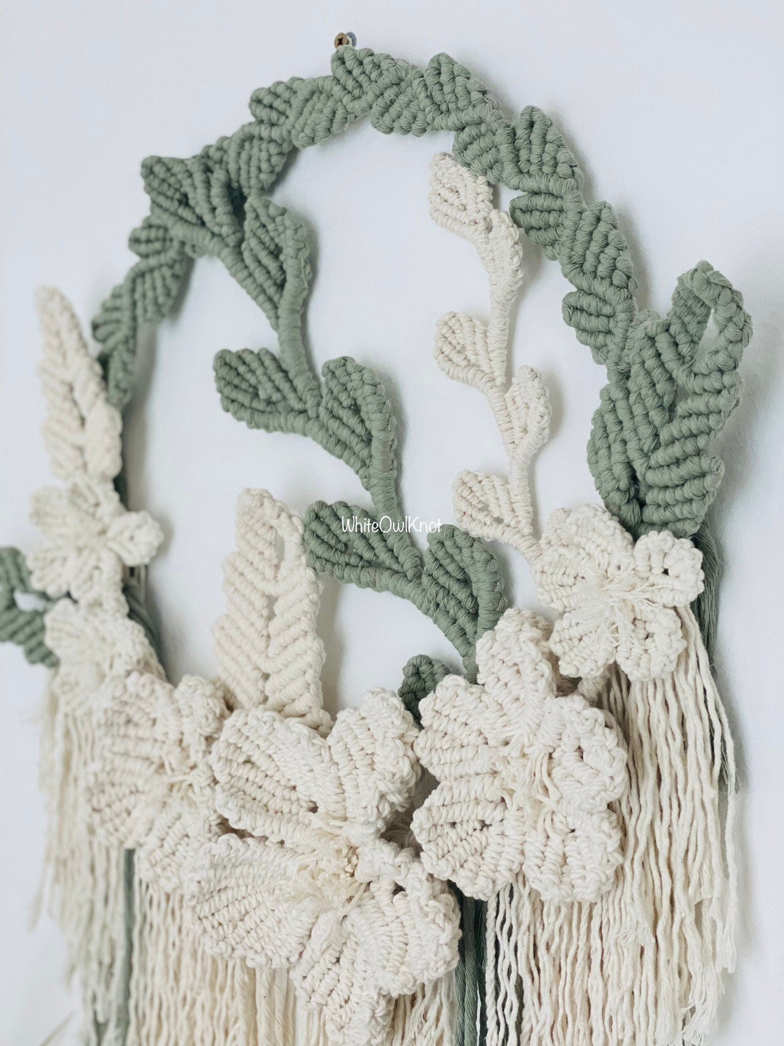 Macrame wall hanging with green and white leaves and flowers on a light background