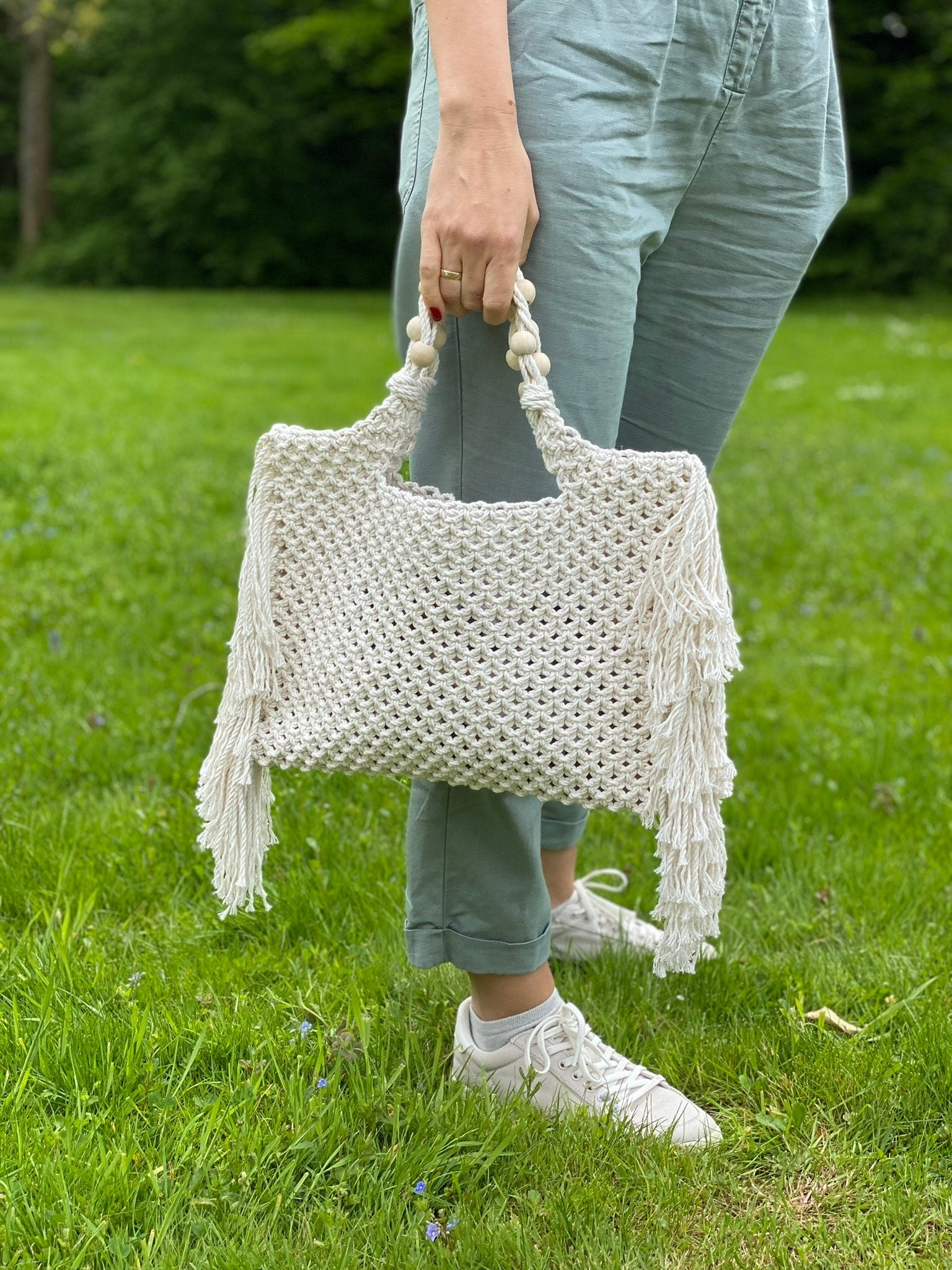 Person holding a white macrame bag with tassels in a grassy outdoor setting