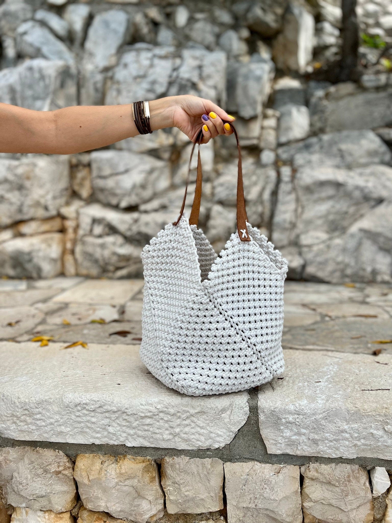 White woven handbag with brown handles held by a person against a stone wall background