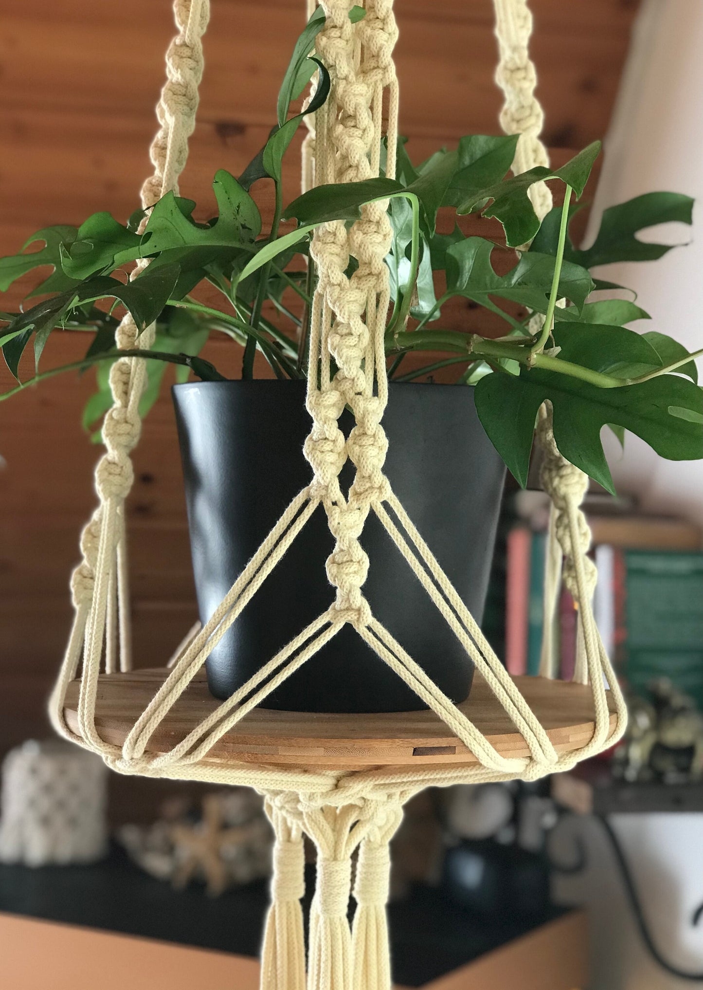 Macrame plant hanger with a potted plant on a wooden shelf against a blurred indoor background.