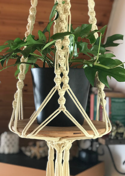 Macrame plant hanger with a potted plant on a wooden shelf against a blurred indoor background.